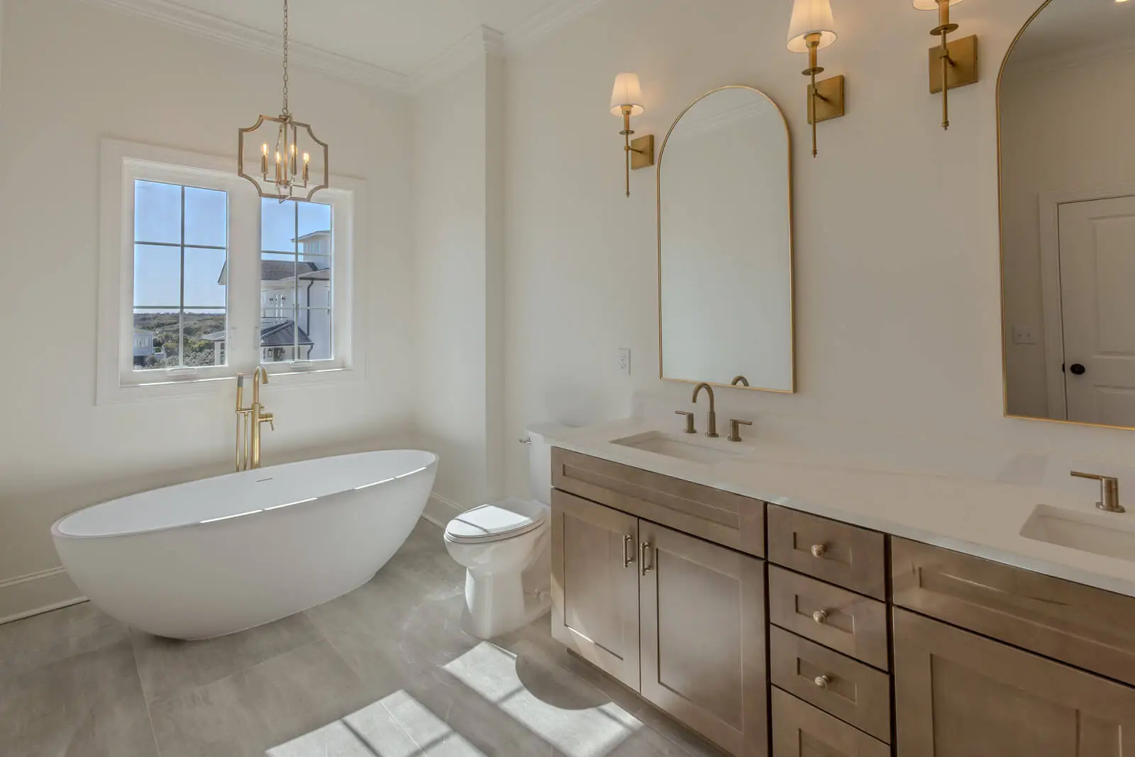 Elegant bathroom with freestanding tub, double vanity, gold fixtures, arched mirrors, and large window with coastal views.