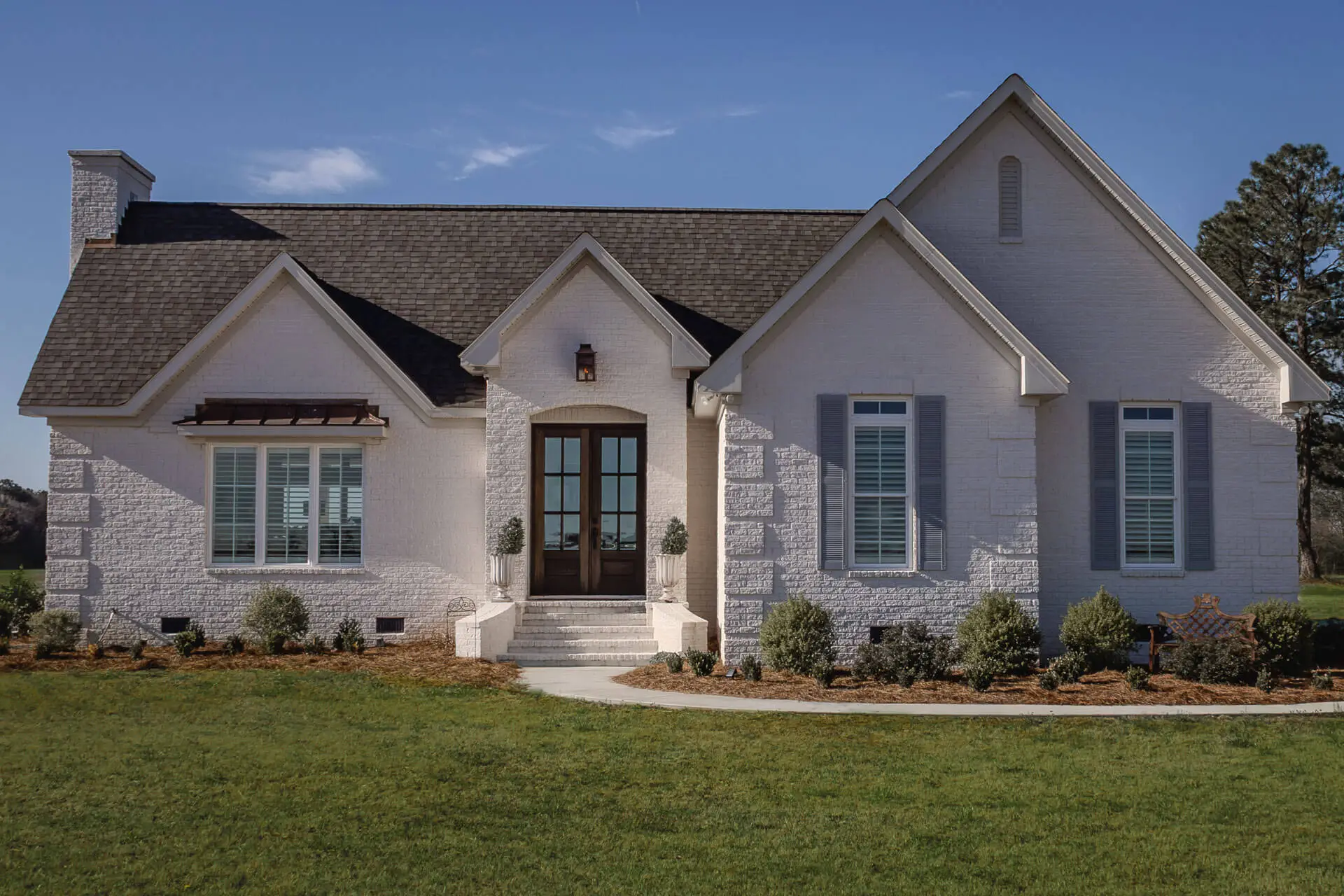 Elegant white brick home with arched double doors, gable rooflines, and shuttered windows framed by neat landscaping.