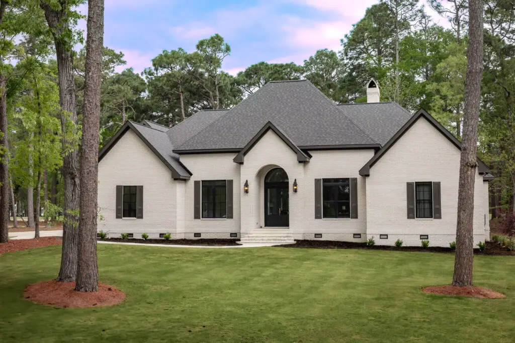 Elegant French Country home with white brick, dark roof, black accents, and manicured lawn amid tall pine trees.