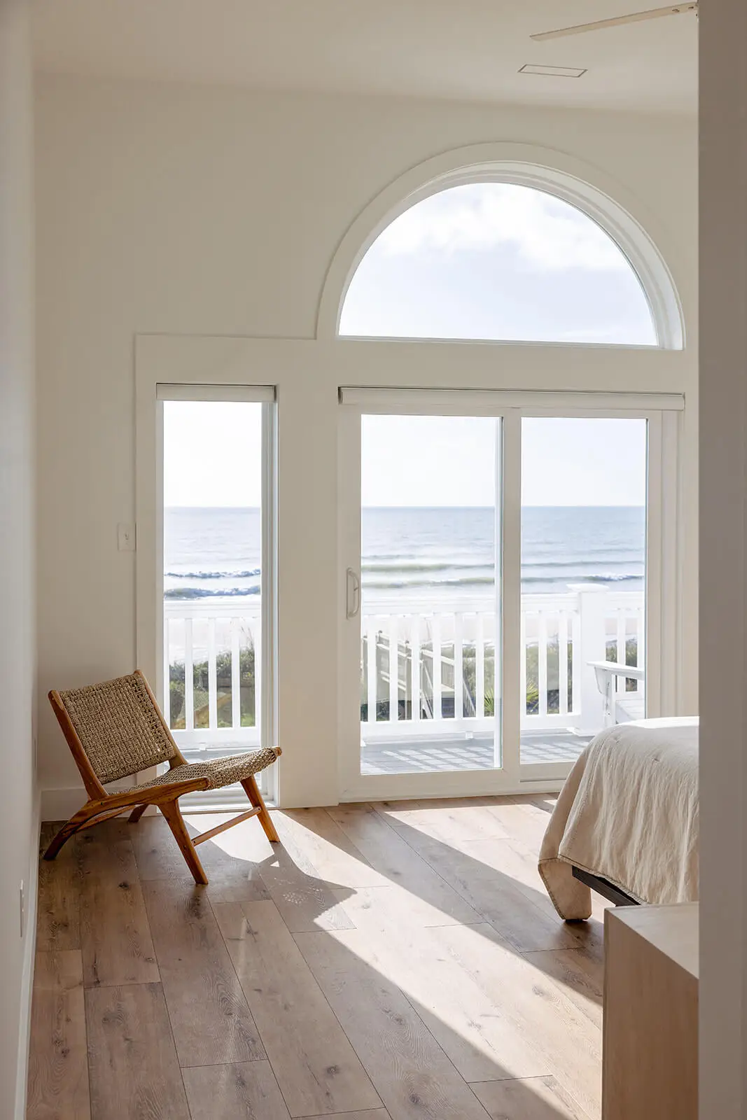 Bright coastal bedroom with arched window, sliding glass doors, ocean view, natural wood floors, and woven accent chair.