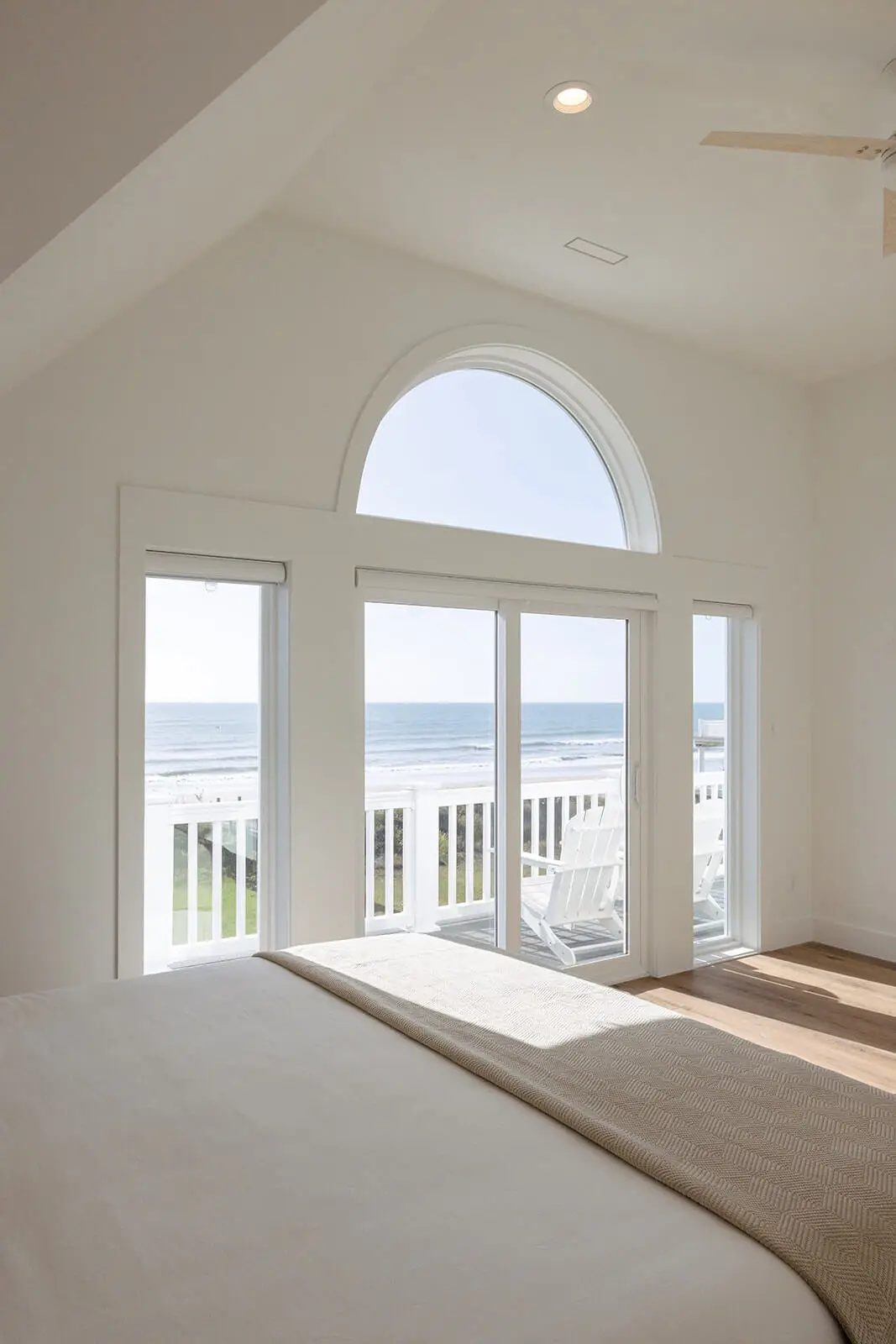 Bright coastal bedroom with arched window, sliding glass doors, and ocean views from a private balcony with Adirondack chairs.